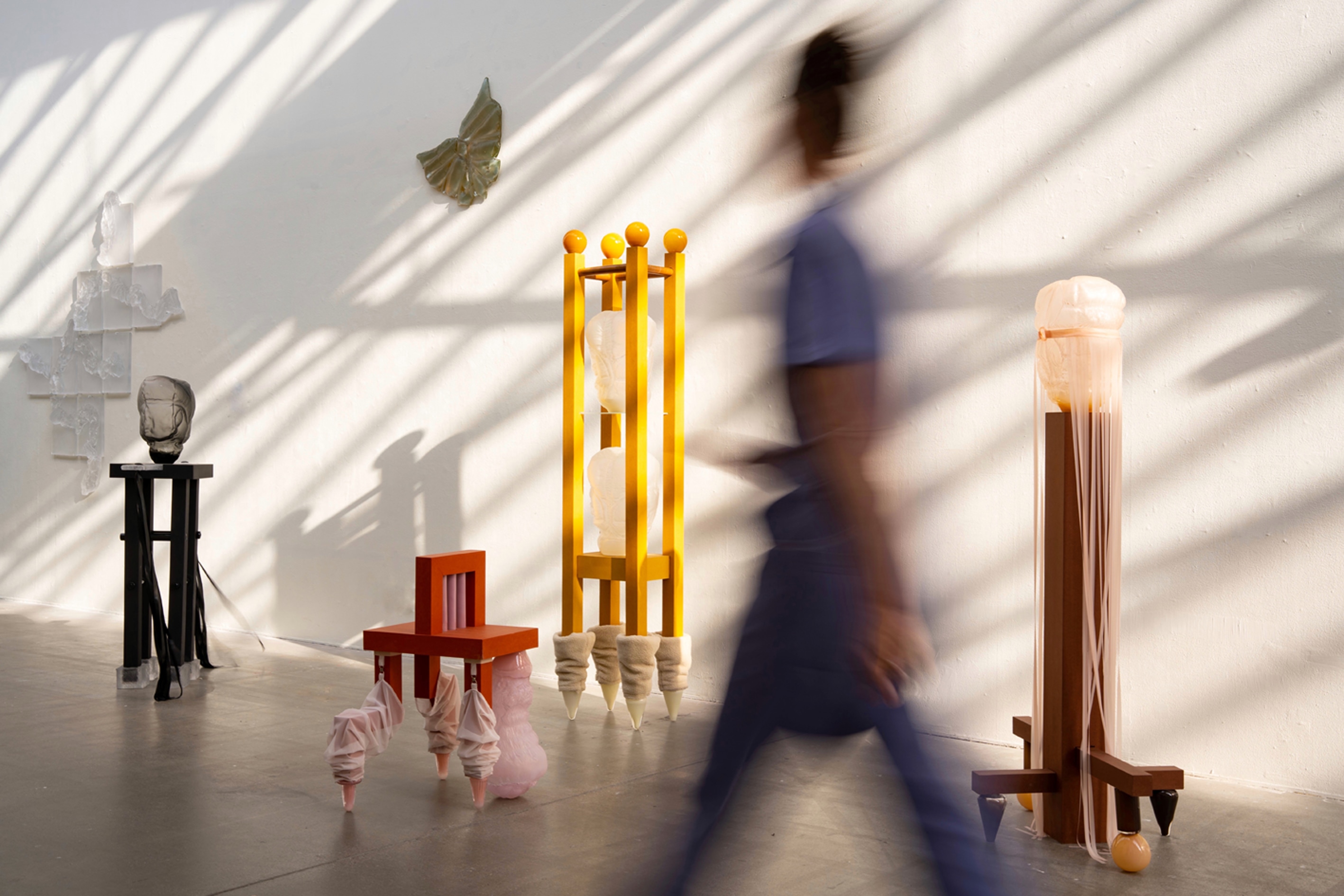 Exhibition view in streaming sunlight: glass forms on furniture-like stands in black, red, yellow and brown, two glass wall pieces, and a motion-blurred person walking past.