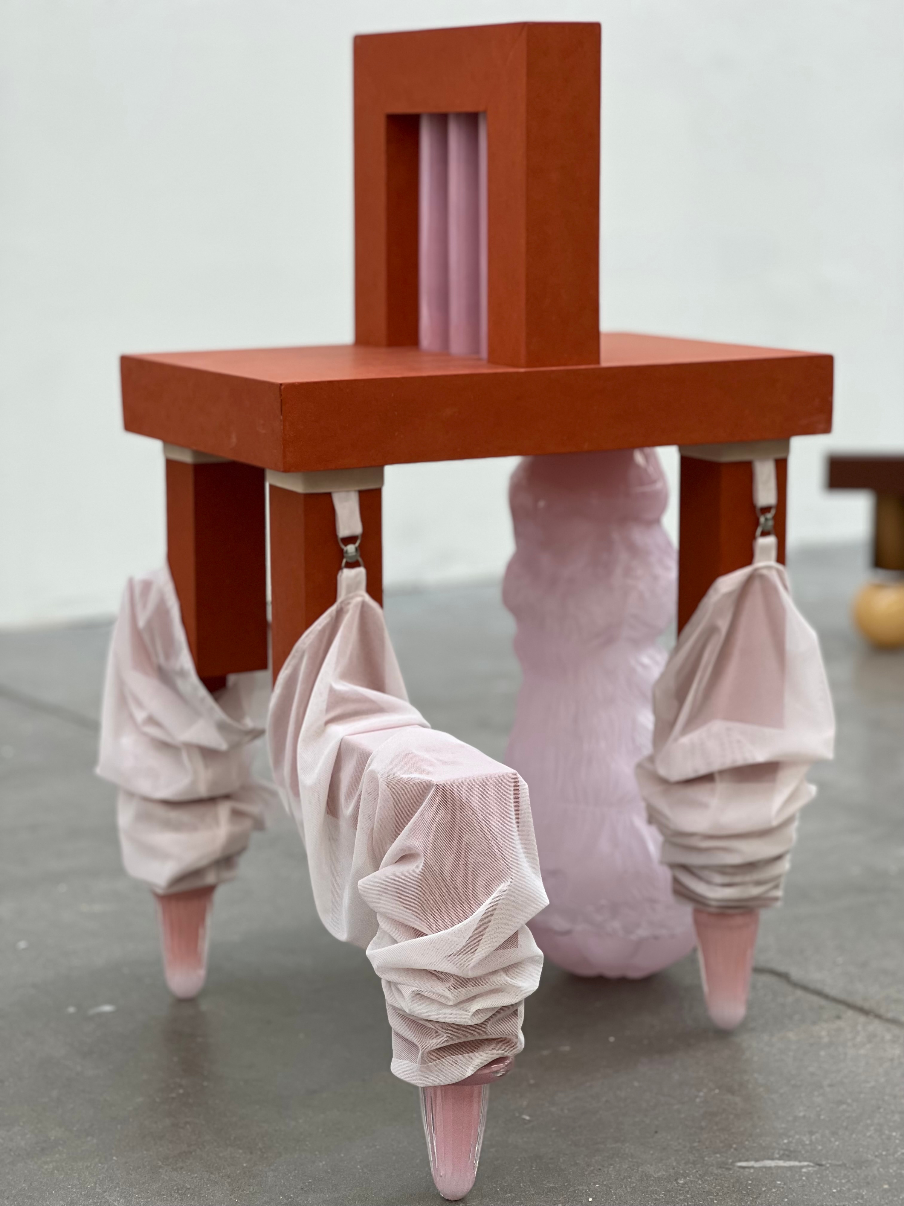 A rust-red chair-like stand with pink glass rods in its backrest, its legs sheathed in gathered pink mesh ending in glass cones, beside a lumpy pale pink glass form under the seat.