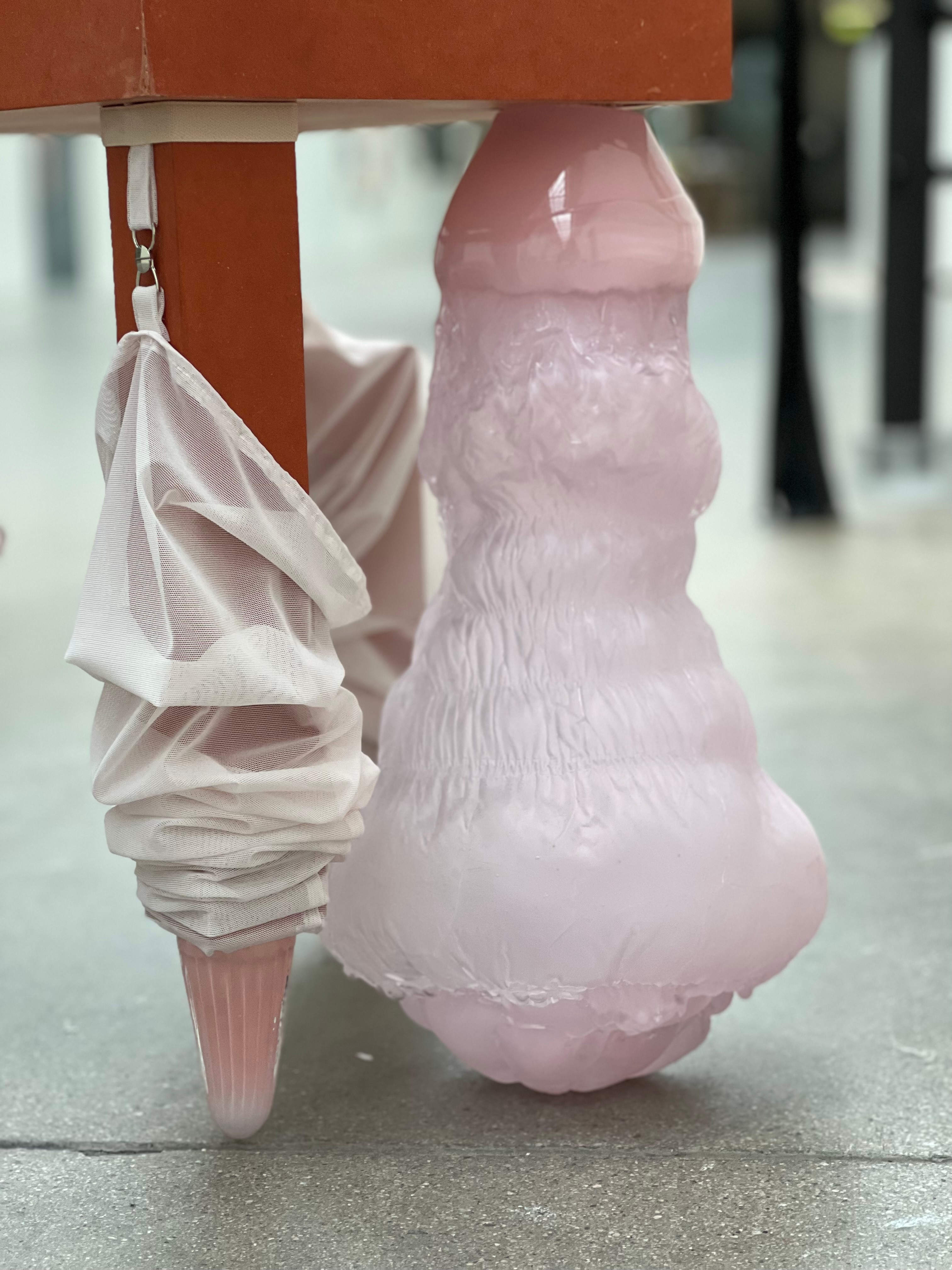 Close-up beneath a rust-red seat: gathered pink mesh hanging from a clip ends in a ribbed pink glass cone, beside a pale pink glass form with a glossy neck and ruched, fur-like texture.