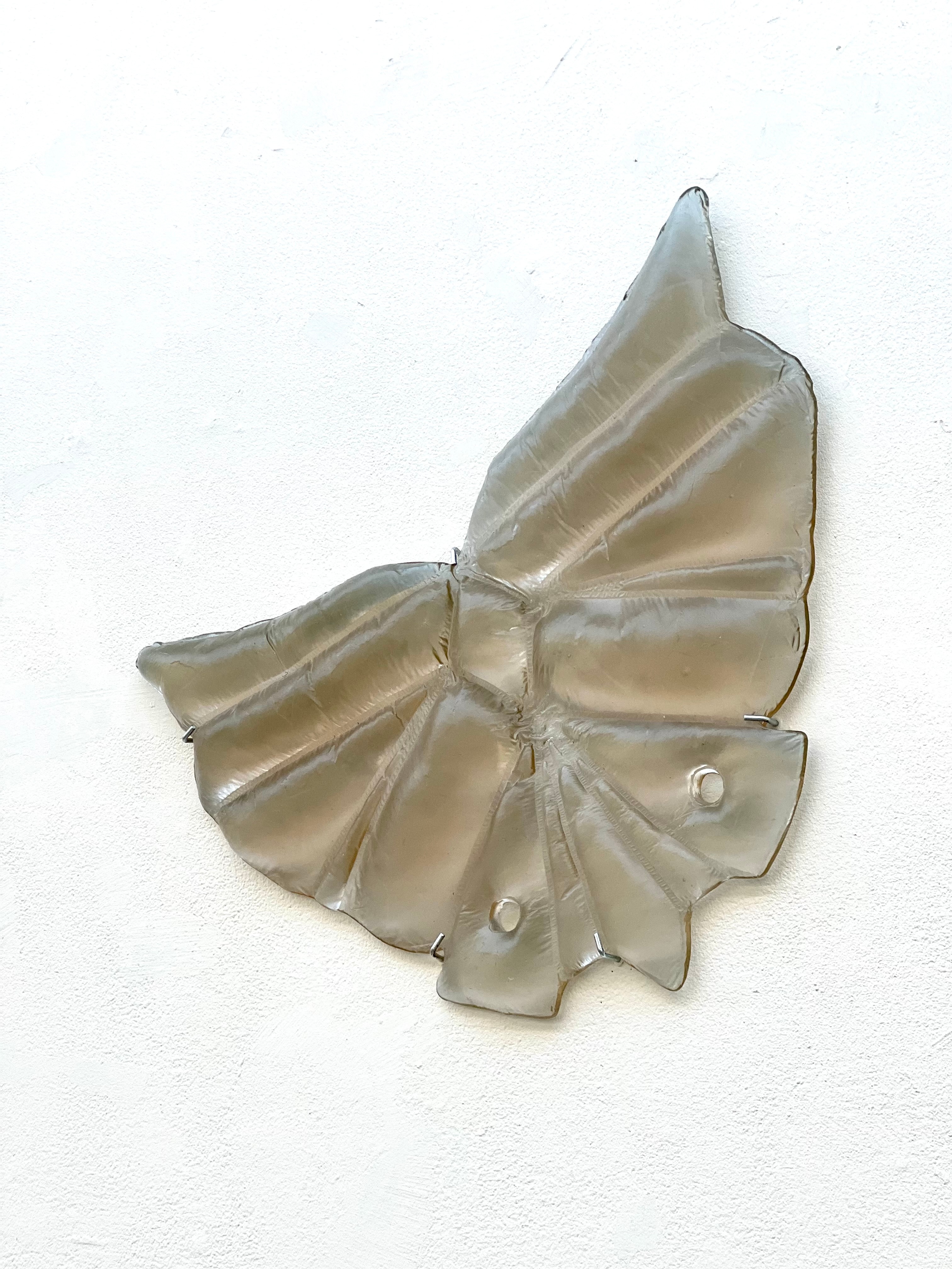 A translucent smoky grey-brown glass wall relief shaped like spread wings, with puffed, stitched-looking panels and two small holes, held to a white wall by metal clips.