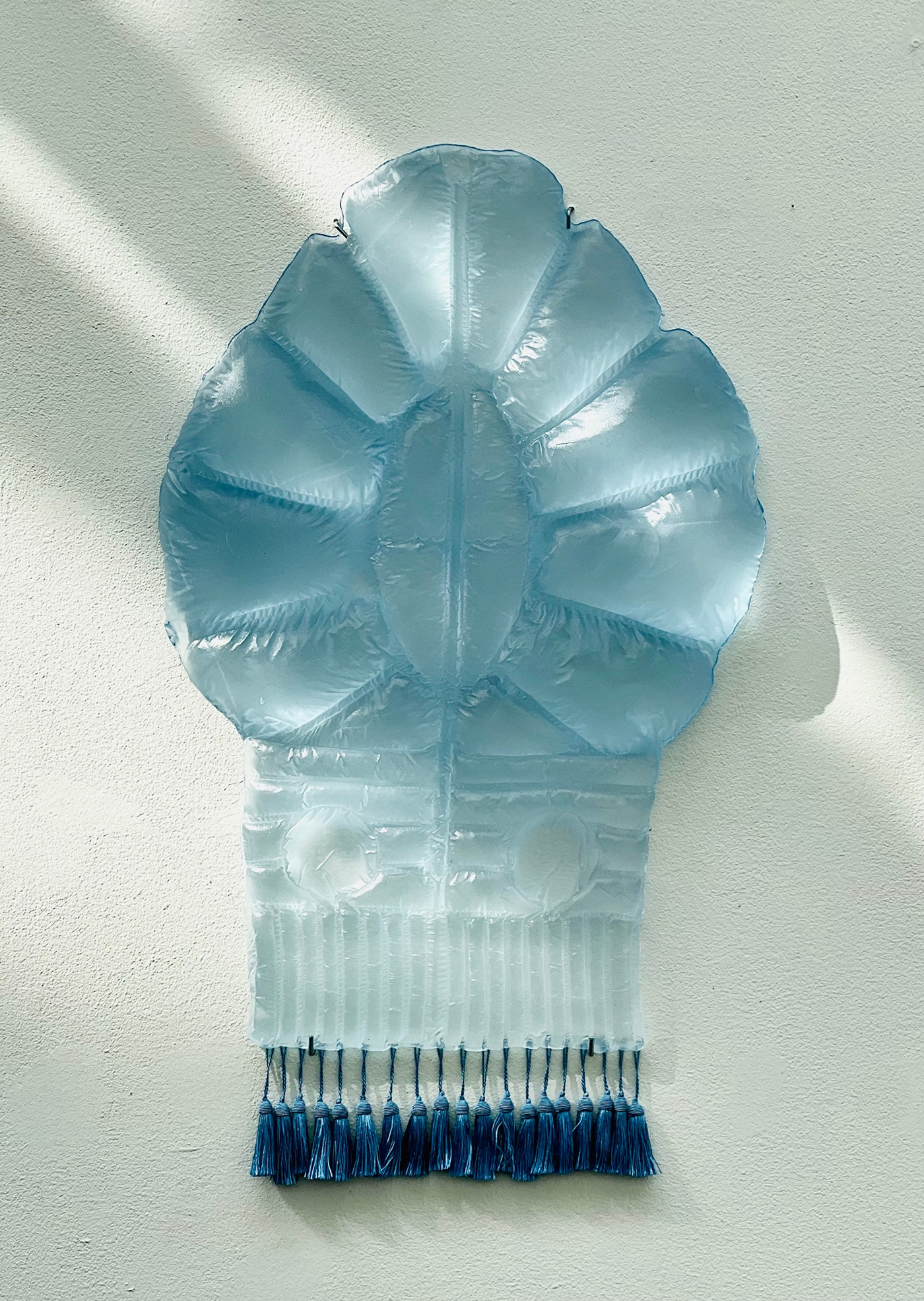 A pale blue glass wall relief with puffed, petal-like panels above a ribbed, quilted lower section, fringed with a row of blue tassels, on a white wall in soft diagonal light.