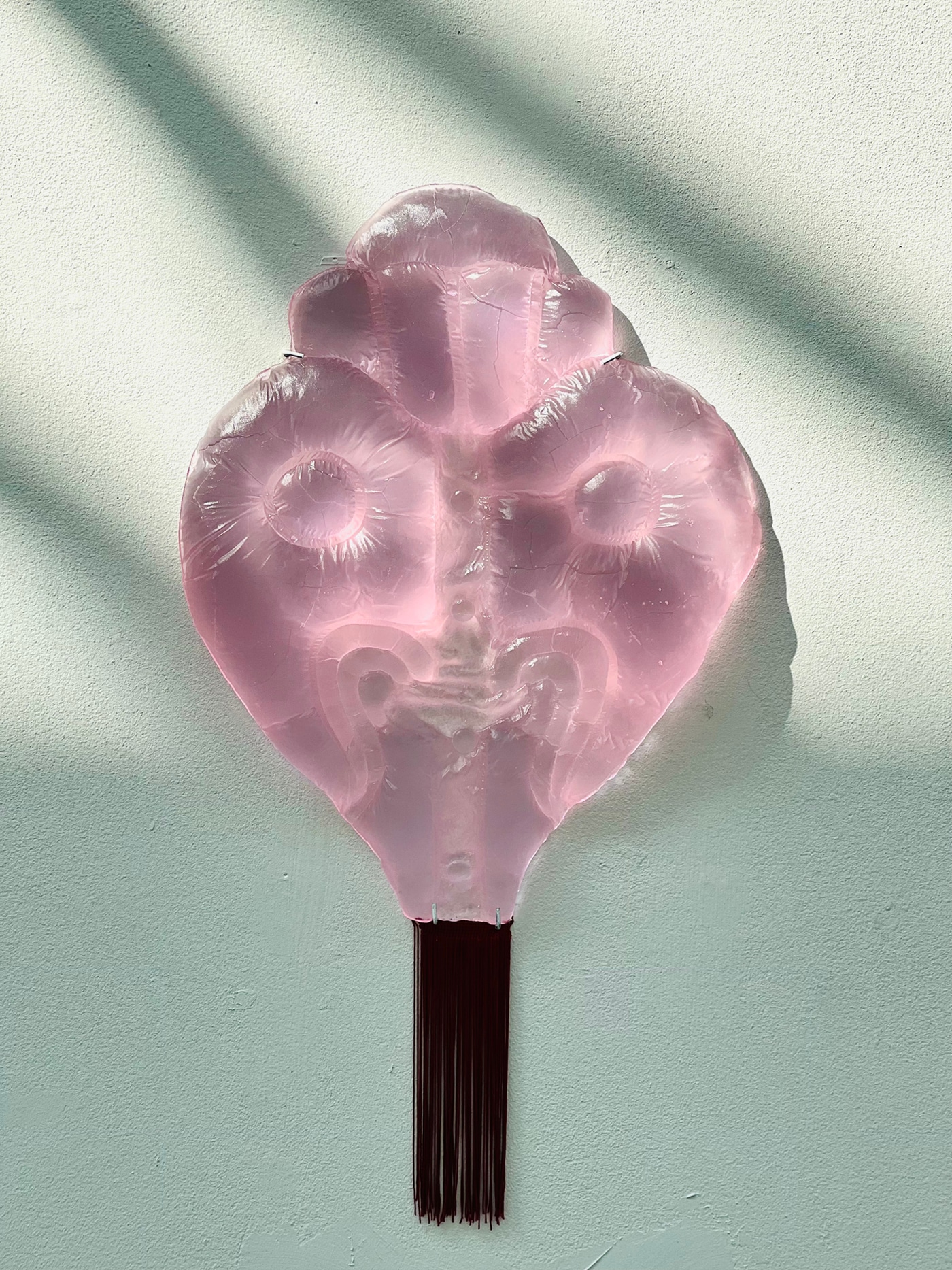 A translucent pink glass wall relief shaped like a face with round, puffed eyes and quilted ridges, a dark red fringe hanging below, on a pale wall in dappled light.
