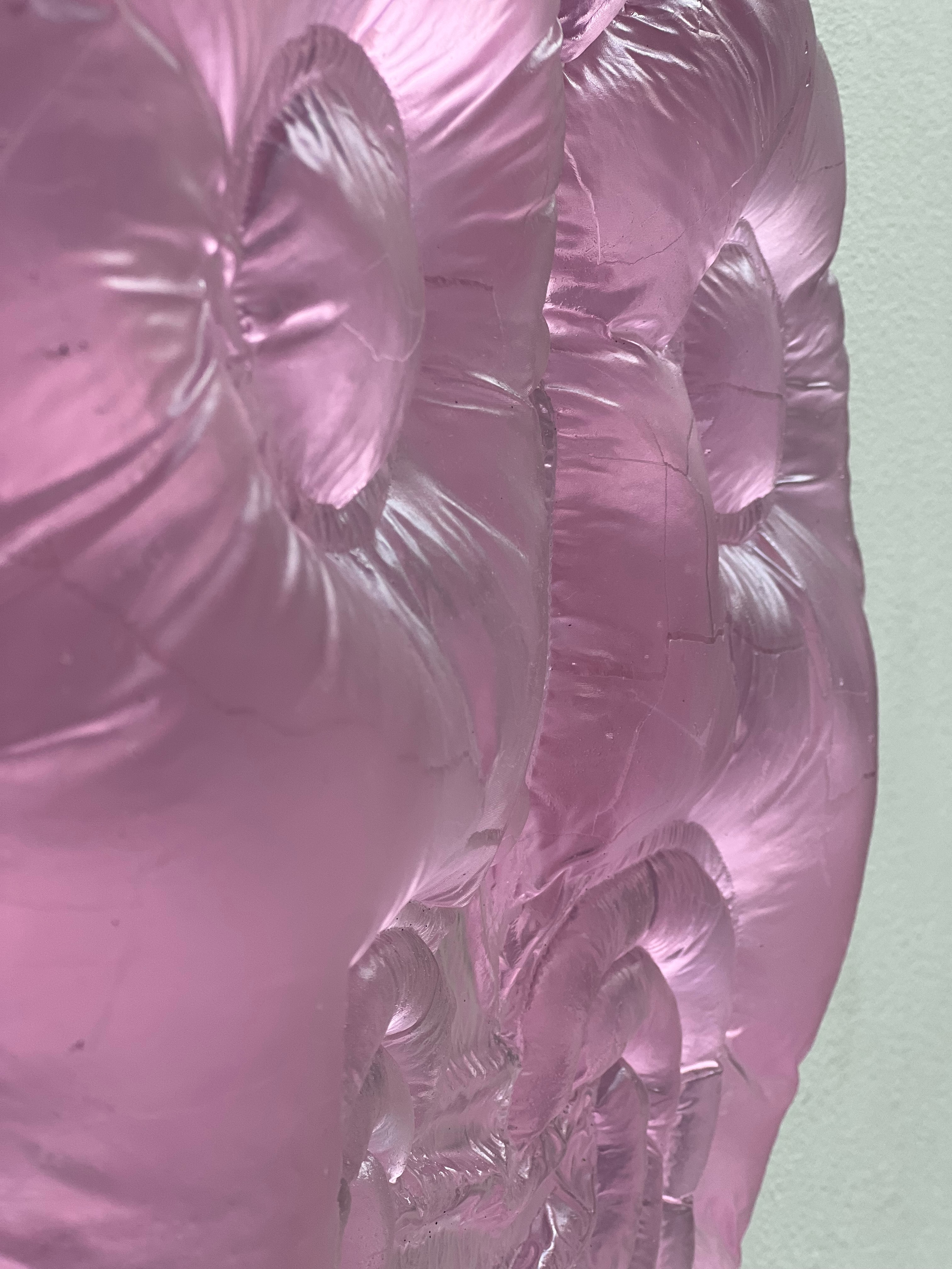 Close-up of glossy pink glass with puffed, quilted swells, fine wrinkles and small cracks, like padded fabric, against a pale wall.