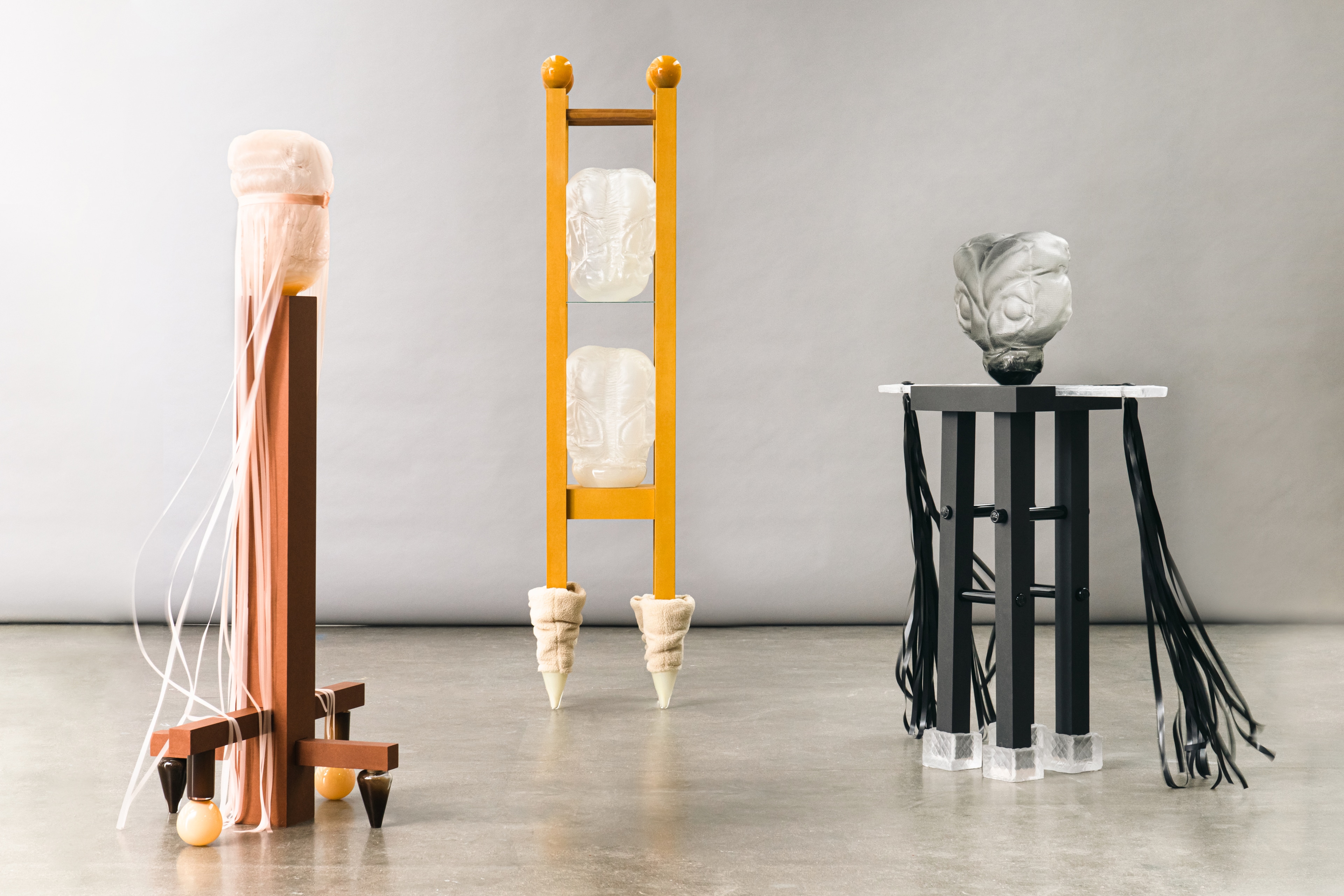 Three of the sculptures side by side on a concrete floor before a grey backdrop: a peach glass form trailing pale ribbons on a rust-brown stand with amber glass feet, two clear glass forms held in a tall yellow ladder-like frame on legs sheathed in beige fabric, and a smoky grey glass head on a black stand hung with black straps.