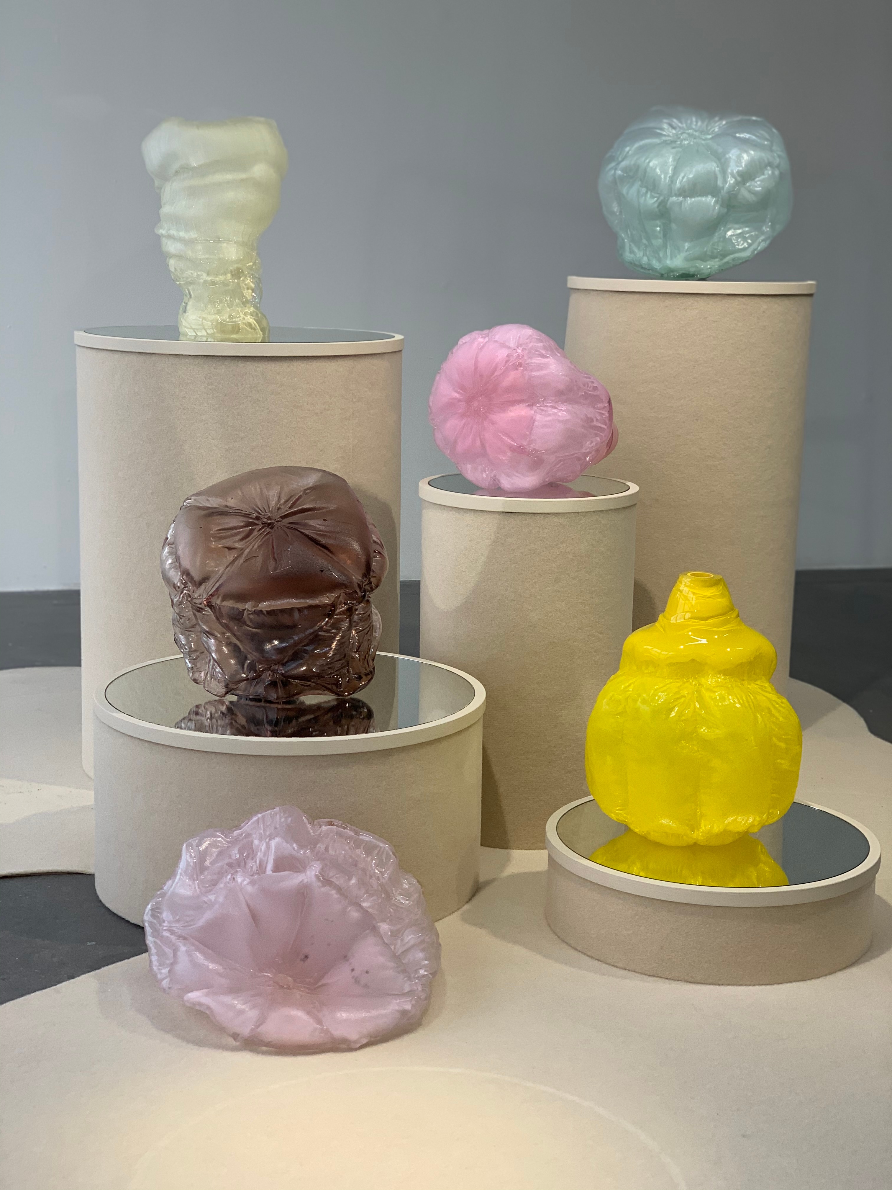 Six quilted, cushion-like glass forms in yellow, pink, aqua, smoky mauve, lilac and pale yellow on mirror-topped, felt-covered round plinths and a cream felt floor in a grey gallery.