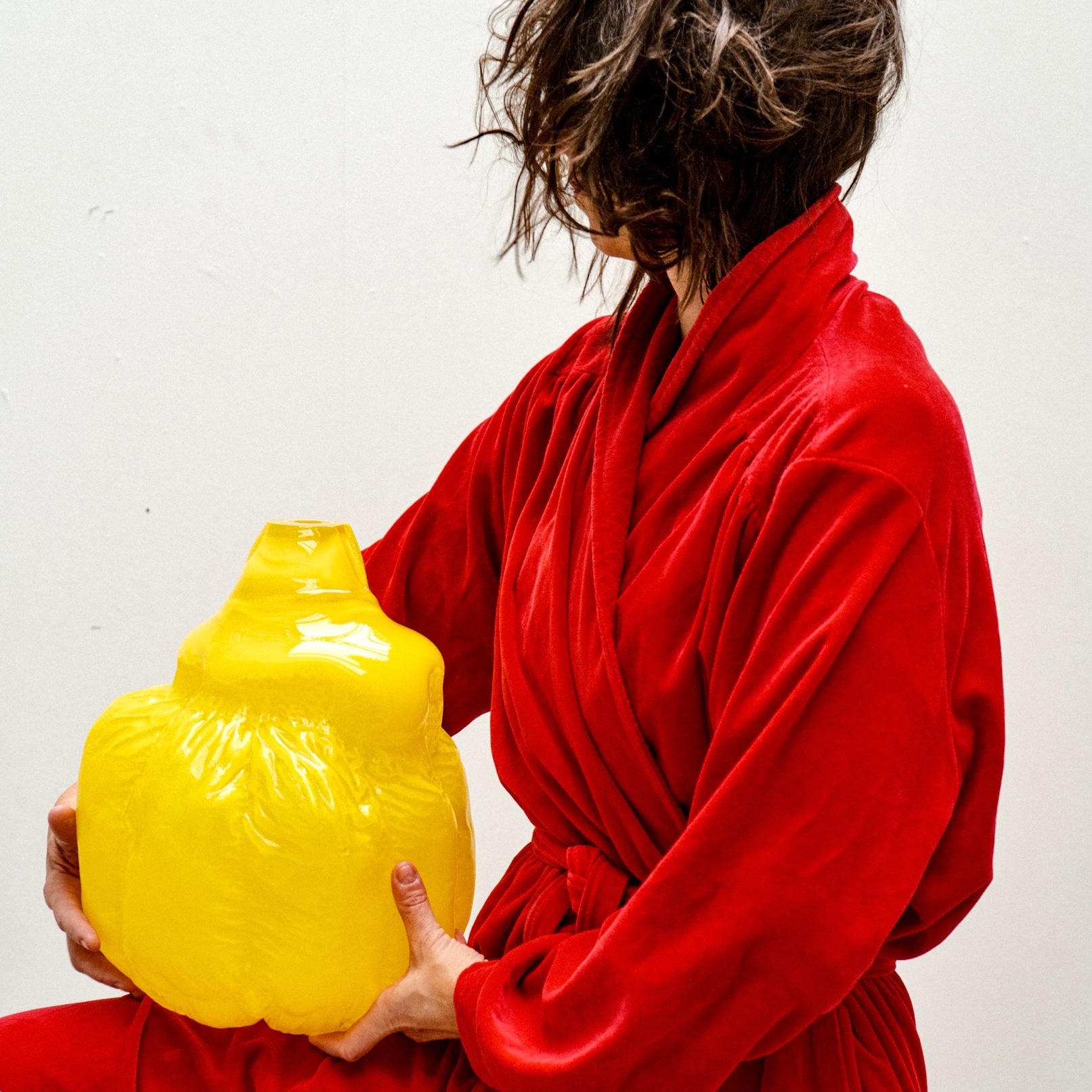 A person in a red velvet robe, face hidden by hair, holds a glossy bright yellow glass vessel with a quilted, pillow-like body against a white wall.