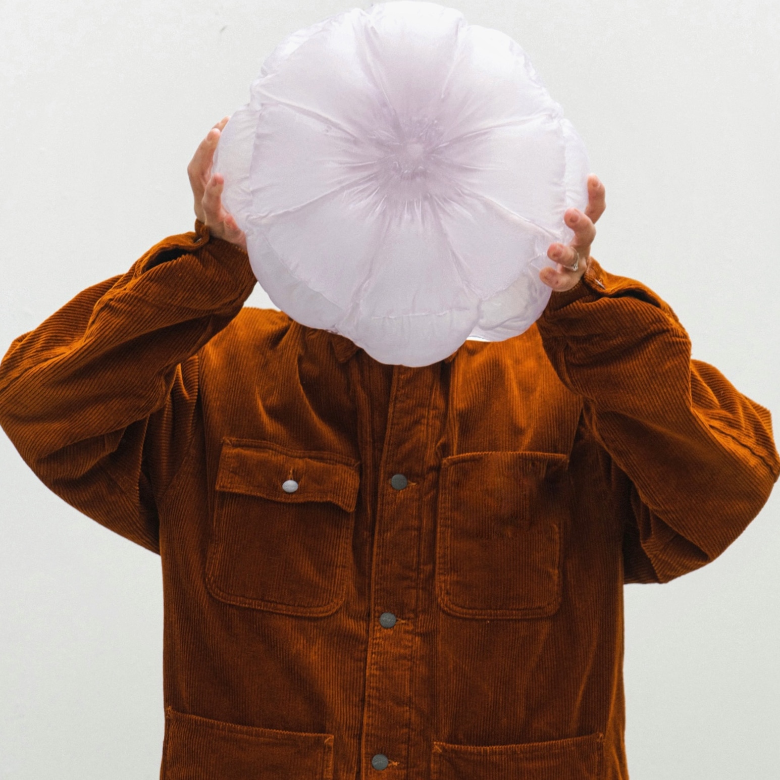 A person in a rust-brown corduroy jacket holds a round, translucent lilac glass form in front of their face; its quilted surface radiates from the centre like a flower.