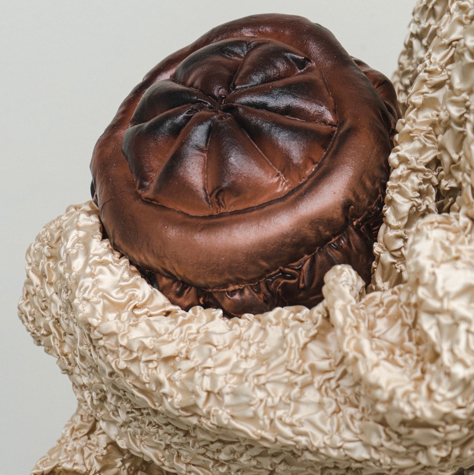 Close-up of a glossy copper-brown glass form with a quilted, cushion-like top, nestled in a fold of crinkled champagne satin against a pale grey backdrop.