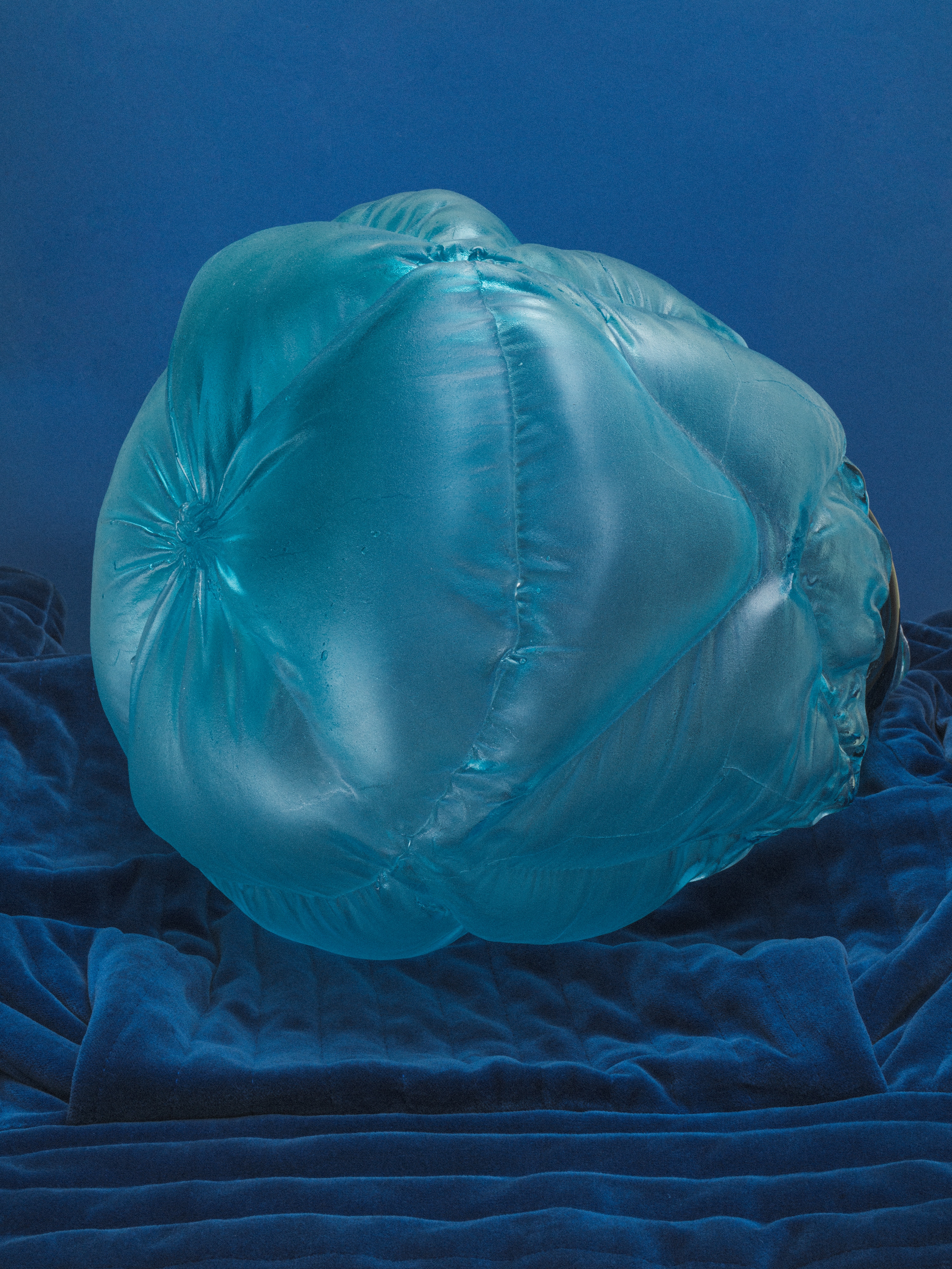 A large translucent aqua glass form, puffed and quilted, its seams gathering at one side, resting on deep blue velvet against a blue backdrop.